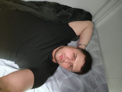 A male individual is depicted lying on his back in an indoor setting within the city of Im. Gastello, Russia. The man has short, dark hair, light facial stubble, and wears a black short-sleeved t-shirt. A silver-colored chain bracelet is present on his left wrist. His left arm is bent, with the forearm positioned near his head. He lies on a light-colored fitted sheet with a repeating dark grey wave-like or dotted pattern. A dark grey blanket or comforter is partially visible behind his head. To the right of the subject, a plain light grey wall with a white horizontal trim piece is visible. The individual's gaze is directed towards the camera.