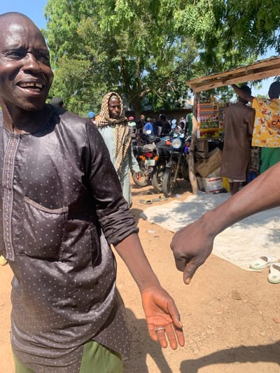 In Project, Nigeria, a man in a dark patterned tunic and green pants stands in the foreground, looking to his left and smiling broadly. His right arm is extended, and a second person's hand is visible in the lower right corner, pointing downwards with a thumb. In the background, other people are gathered around motorcycles and a market stall under a tree. The ground is sandy, and the scene is illuminated by bright sunlight.