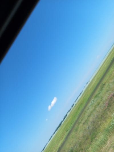 The image shows a wide, bright blue sky with a single white cloud. The sky occupies the majority of the frame. A dark, shadowy object, possibly the edge of a structure or vehicle, is visible in the top left corner, angled diagonally across the sky. Below the sky, a blurred landscape of green fields and a dirt path stretches towards the horizon. Small, indistinct patches of pink and white flowers are scattered in the grassy areas. The scene suggests an open, rural environment under a clear sky. The location in Бөрлі, Kazakhstan, indicates a likely agricultural or natural setting within that region. There are no people or animals visible in the image.