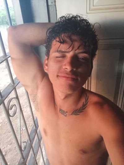 A young adult male with curly, dark hair is pictured from the chest up. He is shirtless and his left arm is raised, bent at the elbow, with his hand resting on the back of his head. He is looking towards the camera with his right eye partially closed, giving a wink. He has a tattoo on his collarbone of what appears to be a laurel wreath with the number "2005" underneath. The background shows a metal grate or bars to the left, suggesting a window or doorway. Behind the bars, outdoor foliage and a sandy ground are visible. To the right of the male, a wall and a door frame are present. The lighting suggests it is daytime. The setting is identified as La Guardia, Venezuela.