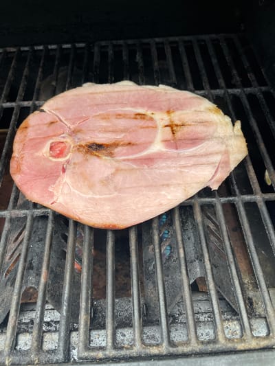 A slice of ham is cooking on a grill. The ham is a pale pink color with visible fat marbling. Grill marks, which are browned and slightly charred, are visible on the surface of the ham. The ham is lying on a metal grill grate, which is dark and appears to be seasoned. Flames are visible through the gaps in the grill grate, indicating that the grill is on and the ham is actively cooking. The background of the image is dark, likely the interior of a grill lid. There are no people or other discernible objects in the frame. The scene is outdoors. The location in Brantingham, United States is not specified in the image.