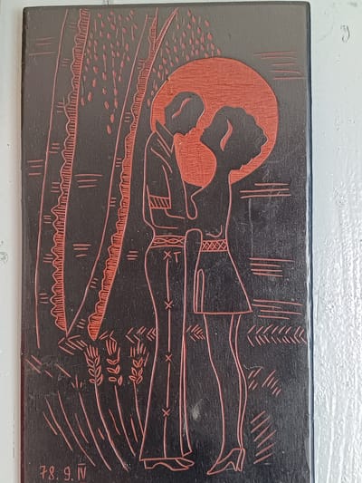 The image depicts a rectangular wooden plaque with a relief carving. The carving is primarily black with red accents. In the center, a male and female figure are embracing. The male figure, on the left, is wearing trousers and a shirt with decorative lines. The female figure, on the right, is wearing a dress with a patterned waistband and high-heeled shoes. Behind them, a large red circle represents a sun or moon. To the left of the figures are stylized tree trunks with rough, serrated edges and lines. Below the figures are carved representations of wheat stalks. In the lower left corner, a date is inscribed: "78. 9. IV". The plaque is displayed against a white, textured wall. The image does not contain any identifiable landmarks or features that would indicate its location in Tebras, Latvia.