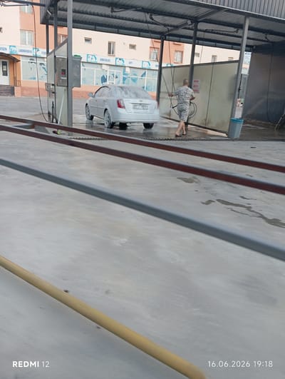 In Bekabad, Uzbekistan, a man in camouflage shorts and a patterned shirt is washing a white Chevrolet sedan at a car wash. He is holding a high-pressure water hose, spraying the vehicle which is partially covered in foam. The car wash is under a metal canopy, with a building visible in the background. The ground is wet concrete. The timestamp in the lower right corner indicates the image was taken on June 16, 2026, at 19:18.