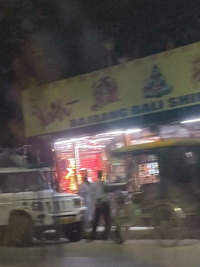 An out-of-focus photograph shows a storefront at night. The sign above reads "BAJRANG BALI SH." with images of deities. The storefront has a closed metal security gate. A white SUV is parked in front, partially obscuring a two-wheeled vehicle. Two men are standing near the vehicles, their faces blurred. The scene appears to be dimly lit, with lights inside the store providing illumination.