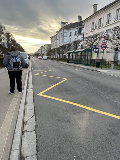 This image depicts a street scene in Serris, France, under an overcast sky. In the foreground, a person stands on a light-colored concrete sidewalk, with their back to the viewer. They are wearing a grey puffer jacket, dark trousers, light-colored sneakers, and a distinctive multi-colored backpack. A grey granite curb separates the sidewalk from a dark asphalt road.

Yellow zigzag lines are painted on the asphalt roadway adjacent to the curb, indicating a specific zone. Further down the street on the right, a transparent green and metal bus stop shelter is visible, accompanied by a black trash bin. Immediately to the right of the bus stop, a pole supports three circular road signs: a triangular warning sign for uneven road, a triangular warning sign for a pedestrian crossing, and a regulatory speed limit sign indicating "30" in a red circle.

A row of pruned deciduous trees lines the street behind the bus stop, fronting a series of multi-story buildings with light-colored facades, multiple windows (some with shutters), and pitched roofs. In the background, the street extends into the distance, with several vehicles (cars and vans) visible on the road, and a traffic light displaying red.