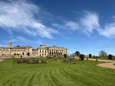The image shows Croome Court, a Palladian mansion located in Croome D'Abitot, Malvern Hills, United Kingdom. The mansion appears to be in a state of disrepair, with visible signs of weathering and deterioration. The building is situated in a large green lawn. A winding, gravel-covered path cuts through the lawn, lined with topiary bushes and small trees. In the background, there's a bright blue sky filled with cirrus clouds. Several people are visible in the image. Some appear to be sitting on the lawn, while others are walking or standing near the mansion. The lawn is neatly mowed, with visible patterns from the lawnmower.