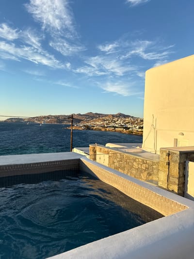 The image depicts a panoramic view of the Mykonian coastline during golden hour. In the foreground, a rectangular outdoor plunge pool or hot tub with deep blue water occupies the lower left, its interior lined with light square tiles and surrounded by white coping. Beyond the pool to the right, a multi-level white building with a flat roof is illuminated by warm sunlight, revealing stone-clad sections and a recessed white door. A dark utility pole with crossarms stands centrally in the midground. The Aegean Sea, a deep blue, stretches across the horizon, dotted with several white sailboats. In the background, a hilly, arid landscape covered with numerous white Cycladic structures defines the city of Mykonos, Greece. The sky above is clear blue with scattered, wispy white cirrus clouds. No individuals are present.