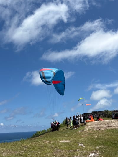The image shows a paragliding site in Benoa, Indonesia. A large blue and red paraglider dominates the foreground against a partly cloudy blue sky. In the distance, near the top right, a group of people stand on a grassy cliff edge. Another paraglider, yellow and orange, is visible in the sky above them. The ocean is visible below the cliff edge. A tandem paragliding team is launching near the edge of the cliff. Another paraglider is in the distance near the water.