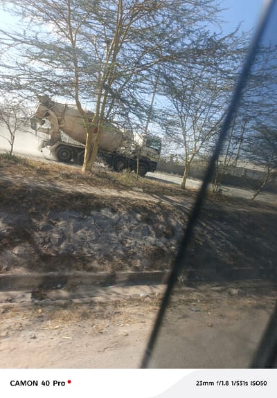 A cement mixer truck drives past trees on a sunny day. Dust rises behind it. A wall of stones lines the road. The photograph is taken from inside a vehicle, as evidenced by the tinted window obscuring the right portion of the frame. The sky is clear.