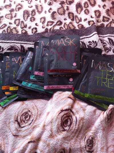 The image shows a pile of dark grey, individually wrapped face masks, each with rose gold lettering and a colored band at the bottom. The masks are arranged on a textured surface that appears to be a pink and beige towel with a brown floral pattern. The background is a leopard-print fabric. The face masks have text that is difficult to read in detail but appears to include brand names and product descriptions. There are no people or any indication of location in Monastirische, Russia.