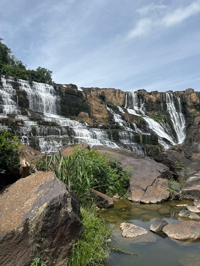 The image shows a wide, multi-tiered waterfall cascading down a rocky cliff face. The water flows over numerous ledges, creating a frothy white spectacle against the dark rock. Lush green vegetation, including tall grasses and trees, grows on the cliff ledges and at the base of the waterfall. In the foreground, large, dark rocks are scattered around a shallow pool of water, which reflects the sky and surrounding landscape. The sky above is partly cloudy with a mix of blue sky and white clouds. The scene is natural and appears to be a scenic location. This waterfall is identified as the Pongour Waterfall, located in Tân Phú, Vietnam. There are no people visible in the image.