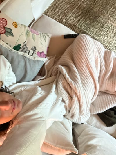 The image depicts an individual resting in a bed or similar soft surface, located in Vila Nova de Cerveira, Portugal. The subject is oriented on their back, partially visible from the mid-chest upwards, with their head supported by multiple pillows. They have dark, wavy hair and are wearing a white cable-knit sweater. The left side of the subject's face, including an eye, nose, and mouth showing a slight smile, is visible.
Surrounding the individual are several pillows: a cream-colored pillow with a floral pattern featuring red, pink, green, and black elements; a grey pillow with a herringbone pattern; and additional plain white or light-colored pillows. A light pink or beige ribbed knit blanket or throw is draped over the lower body and extends to the right, partially obscuring a dark green or brown fabric item. A beige woven rug is visible in the upper right background. A black rectangular object, consistent with a remote control, rests on a light-colored surface in the upper center of the frame. The scene is illuminated by soft, warm light.