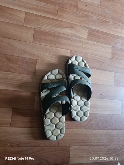 A pair of olive green and tan hexagonal patterned flip-flops are lying on a light brown wooden floor. The flip-flops are positioned on the lower half of the image, with the left flip-flop slightly above and to the left of the right flip-flop. The floorboards are oriented diagonally from the top left to the bottom right. The image was taken in Omyak, Russia.