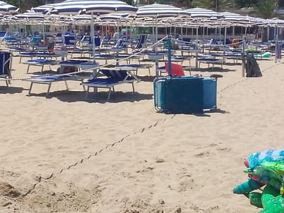 The image depicts a sunny day at a beach in Valle Castellana, Italy. The scene is filled with rows of blue and white striped umbrellas and lounge chairs, set up on a sandy shore. In the foreground, a blue plastic container is visible, along with part of a colorful inflatable toy, possibly a crocodile. The sand shows faint tracks, suggesting recent movement. In the background, more umbrellas and chairs extend towards a building with a red roof. The overall impression is one of a well-established beach resort ready for visitors, with a clear sky and warm weather implied by the presence of beach amenities. No people are clearly depicted, though figures are faintly visible in the distance. There is no visible text in the image.