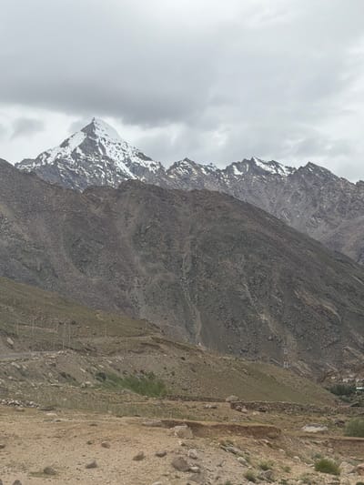The image depicts a mountainous landscape near Reru, India, under a cloudy sky. A prominent snow-capped peak dominates the background, with jagged, rocky mountains extending across the horizon. The middle ground features steep, barren slopes covered in scree and sparse vegetation. A winding road can be seen traversing the lower slopes, with a few utility poles lining its path. The foreground consists of a rocky, arid terrain with scattered stones and dry grass. In the far distance, a small cluster of buildings, likely a village, is visible nestled in a valley between the mountains. The scene conveys a sense of rugged, remote wilderness.