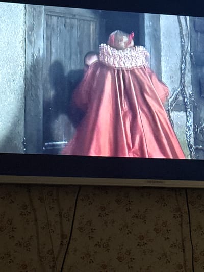 The image shows a television screen displaying a scene of a person in a red gown lying on a wooden platform or stairs. The person is positioned head-down with their head facing the top of the screen. Their fair hair is visible. The gown has a ruffled neckline. The platform or stairs appears to be made of rough-hewn wood and is set against a background that looks like stone. The word "HYUNDAI" is partially visible along the right edge of the screen. Adjacent to the screen is a floral patterned surface. The image does not provide any information to confirm or deny the location of the image in Скалба Снт, Russia.