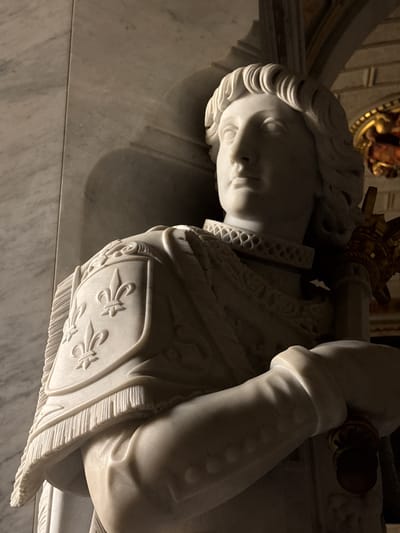 The image shows a marble statue of a male figure, likely a king or a saint, dressed in ornate armor. The statue is positioned indoors, with marble walls and a hint of golden architectural decoration in the background. The figure is depicted from the chest up, with a regal expression and elaborate detailing on the armor, including fleur-de-lis symbols on a shield. The statue is lit by a spotlight from the left, casting shadows that emphasize the texture and form of the marble. This statue is located in the Royal Pantheon of El Escorial Monastery, in San Lorenzo de El Escorial, Spain, specifically within the Basilica of the Monastery, which houses the tombs of Spanish royalty.
