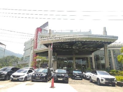The image depicts the exterior of a multi-story hotel building and its associated parking area on a clear, sunny day, located in the city of La Roche-en-Ardenne, Belgium. The modern hotel structure features a prominent red vertical facade element on its left side and a rooftop sign indicating "Hotel Horison Puncak." The building's front is composed of light-colored panels and extensive glass windows. A large, geometrically patterned canopy extends over the main entrance, beneath which a red and white flag is displayed on a pole.
In the foreground, an asphalt or paved parking area contains approximately eight passenger vehicles. These include various models such as a silver Honda hatchback, a black Nissan X-Trail SUV, a black Honda SUV, and multiple white SUVs/crossovers, including a white Hyundai Creta prominently parked in the right foreground. An orange traffic cone is placed on the pavement near the center of the foreground.
The background on the left reveals lush green mountains. Overhead, multiple utility lines traverse the bright, clear sky. No individuals are visible or identifiable within the scene.