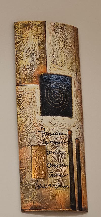 The image shows a textured, abstract artwork mounted on a light-colored wall. The artwork is rectangular and oriented vertically. It features a mosaic-like pattern of squares and rectangles with varied textures and colors, primarily in shades of beige, gold, brown, and black. A prominent black square in the upper center contains a concentric spiral design. Below this, black script is written over the textured surface. There are also two dark, vertical rectangular elements on the right side of the artwork. The artwork's textured surface creates a play of light and shadow, enhancing its abstract quality. The artwork's location in Dogato, Italy, is not determinable from the provided image.