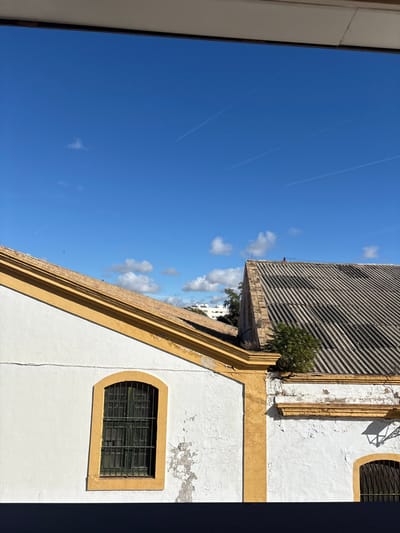 The image displays an urban rooftop scene in El Puerto de Santa María, Spain, under a clear, bright blue sky. Scattered white cumulus clouds are visible, along with three distinct white aircraft contrails traversing the upper portion of the sky. The immediate foreground is bisected by a dark horizontal bar, indicative of a window frame or balcony railing.
Two building structures dominate the middle ground. The building on the left has white stucco walls, exhibiting areas of peeling paint and dirt accumulation. An ochre-yellow trim outlines its gabled roof and frames a recessed, arched window fitted with dark green metal security grilles. The gabled roof is covered with weathered, light brown terracotta tiles.
The building on the right features a steeply pitched, corrugated roof, dark grey in color, showing significant weathering, discoloration, and streaking. A small, bushy green plant grows from a crevice on the white wall where it meets the roofline. This building's white walls also display peeling paint and are bordered by ochre-yellow trim. A portion of a second arched window with grilles is visible on the far right. In the distant background, beyond the immediate rooftops, white residential buildings are partially discernible, interspersed with green foliage. No individuals or active interactions are observed.