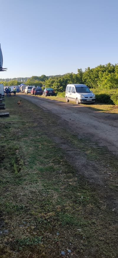 The image depicts an outdoor scene in Bezpalivka, Ukraine, featuring a line of parked vehicles along a dirt path beside a grassy area. The sky is clear and blue, with lush green trees bordering the path on the right side of the frame. Strong shadows cast by the vehicles suggest it is daytime, possibly morning or late afternoon.
The most prominent object is a silver minivan, likely a Renault Kangoo, parked on the dirt path in the midground. Its license plate, "HB 1980 ЕВ", is clearly visible and consistent with Ukrainian registration. Further down the path, several other cars of various colors are parked in a row.
In the foreground, a small, light brown dog is seen walking on the dry grassy terrain. Further into the background, near the end of the line of cars, a group of people is visible, some standing near what appear to be tents or makeshift structures. On the far left edge of the image, part of a larger, dark vehicle (possibly a truck or trailer) is partially visible.
The scene suggests an outdoor gathering or temporary parking area, possibly for an event such as a market, festival, or local meeting, given the informal setup, multiple parked cars, and presence of people and what might be temporary structures.