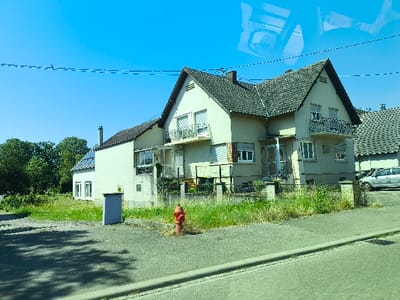 A house features a tan facade, topped with a dark gray roof. A balcony extends from the second story. A small red fire hydrant is positioned in front of overgrown grass along the sidewalk. A grey metal box is visible closer to the house. Nearby, a parked silver car sits by the neighbor's house. The sky is clear and blue.