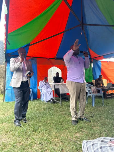 The image, set in Kiganjo, Kenya, captures an outdoor gathering under a vibrant, multi-colored tent with panels of red, blue, and green fabric, supported by metal poles. The ground beneath is covered in lush green grass.

In the central foreground, a dark-skinned man with a clean-shaven head stands, holding a microphone in his left hand and raising his right hand in a dynamic gesture, suggesting he is speaking, preaching, or singing. He is dressed in a light purple long-sleeved shirt and khaki trousers. To his left, another man in a light grey blazer, white shirt, and dark trousers is also standing, holding what appears to be a book or tablet. He wears glasses and looks towards the speaker.

Several other men are visible in the background, mostly seated on light blue plastic chairs. One man in a dark top is seated behind a table with a laptop, seemingly engaged with it. A small framed picture, likely a photograph, is also on this table. Next to him, a man dressed in a white robe or religious garment is seated behind another table. Further back, another man in a brown uniform or suit is seated. The lower body and legs of a person in a plaid skirt are partially visible on the far right.

The bright, natural lighting suggests it is daytime, indicating fair weather. The scene depicts what appears to be a religious service, public address, or community event, with attendees and speakers gathered under the temporary shelter of the tent. No discernible text is visible in the image.