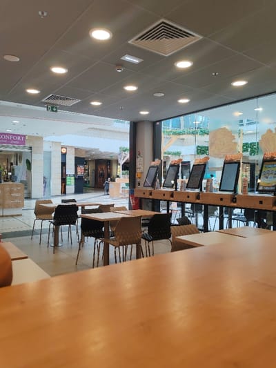 The image depicts the interior of a quick-service restaurant or food court area located within a large shopping center in Fontenay-sous-Bois, France.

The foreground is dominated by a light-colored wooden tabletop. In the midground, several square wooden tables are arranged with a mix of light brown/tan and black textured chairs. To the right, a long, light-colored wooden counter-height structure holds six self-service touchscreen ordering kiosks, each displaying promotional graphics that appear to feature food items resembling tacos or wraps. A partially visible red beverage dispenser is located near the kiosks. Behind this counter, a large glass wall separates the establishment from an outdoor-facing or atrium-like area, revealing distant building facades and greenery.

The background shows a wide, well-lit mall corridor with tiled flooring and multiple retail storefronts, one of which partially displays the word "CONFORT". A few indistinct individuals are visible in the distant mall common areas, walking or standing, but none are clearly interacting within the immediate establishment.

The ceiling consists of a grid of light-colored acoustic tiles, illuminated by numerous recessed circular lights. Square ventilation grilles, a rectangular light fixture, a small round detector, and a green exit sign are also mounted on the ceiling. The scene appears to be in a quiet period, with no active customer interactions observed within the restaurant's seating or ordering areas.