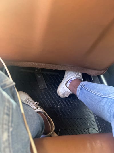Two pairs of legs with shoes are visible in the image. One person is wearing blue jeans and white sneakers with purple-striped socks. The other person is wearing denim jeans and light-colored sneakers. They are inside a vehicle, with the floor covered by a black textured mat. The brown interior of the car, likely a seat, is visible in the upper portion of the image. There are no discernible landmarks or indicators to suggest the location is Yasen, Bulgaria.