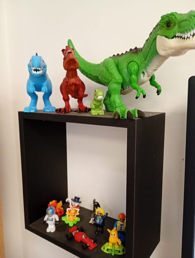 A slightly high-angle indoor shot captures a display of toys on a black shelving unit against a plain white wall.
On the top surface of the black shelf, four dinosaur toys are arranged. From left to right, there is a medium-sized bright blue dinosaur, a medium-sized reddish-brown dinosaur, and a small light green dinosaur. Dominating the right side of the shelf is a larger, detailed green dinosaur with a beige belly and white teeth, facing right.
Below, inside the square opening of the black shelving unit, several smaller figures and toys are displayed. These include a white astronaut figure with a blue helmet on the left, a red race car toy in the foreground, and various other small figures. Among them are a clown-like figure with a top hat, a yellow animal on a green base, a figure wearing a blue jacket and yellow hair, and another figure with an orange hard hat. To the far right, a small yellow creature on a green base is visible.
A dark, rectangular object, possibly part of a monitor stand or another piece of furniture, is partially visible on the right side of the image, adjacent to the black shelving unit. The lighting is soft and even, suggesting an indoor setting without direct sunlight. There is no visible text or specific activity taking place, just a static arrangement of toys.