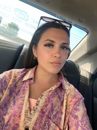 A young woman with long dark hair and false eyelashes is sitting in the back seat of a vehicle. She is wearing a snakeskin-patterned button-down shirt in shades of pink, purple, and beige, over a peach-colored tank top. Her sunglasses are resting on top of her head. The interior of the car is visible behind her, including a black leather seat and the rear window which has text printed on it. The scene suggests she is traveling within the city of Dzhalgan, Russia, as indicated by the presence of the vehicle and typical urban background elements visible through the window.