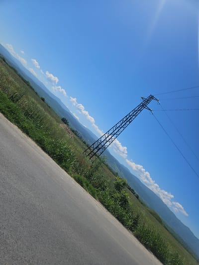 The image shows a rural scene in Yoakim Gruevo, Bulgaria. A power line tower stands in a field of tall grass next to an asphalt road. In the background, rolling hills are visible under a blue sky with scattered clouds. The sun is bright. There are no people visible.