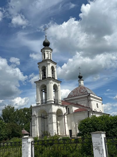 The image shows a white stone church with a tall bell tower and a dome topped with a cross, set against a blue sky with fluffy white clouds. The church has multiple arched windows and decorative architectural elements. In the foreground, there is a black metal fence and a white stone pillar. Lush green trees and bushes surround the church, with a small red-roofed structure visible behind the fence on the left. The overall scene depicts a serene religious building in a verdant setting. This church is likely located in the village of Ерино, Russia, as indicated by the prompt.