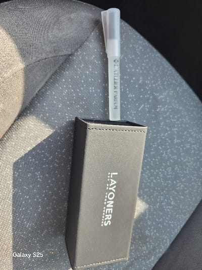 The image depicts a black rectangular container holding a transparent, pen-shaped atomizer on a grey, textured fabric surface, likely a car seat, within Novo Delchevo, Bulgaria. The container, positioned vertically with its longer side parallel to the camera's frame, features the silver text "LAYONERS" and "LAY ON YOUR HORSE" printed on its visible face. The atomizer, inserted into the top of the container, is clear with a translucent white cap and displays the vertical text "L'ATELIER PARFUM." The grey fabric surface exhibits a patterned weave with small white dots and visible stitching. Strong directional lighting from the left casts a prominent shadow of the container onto the fabric. A "Galaxy S25" watermark is present in the lower-left corner. No individuals or active interactions are visible in the scene.