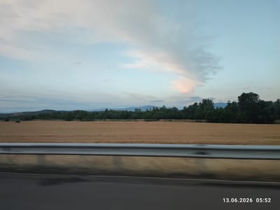 An outdoor, wide-angle, eye-level, daytime shot captures a rural roadside scene. In the foreground, a dark asphalt road transitions into a reflective metal guardrail. Beyond the guardrail, a vast field of golden crops stretches horizontally across the frame, bordered by a dense line of dark green trees. In the distance, rolling hills and mountains are visible under a pale blue sky dotted with wispy, soft clouds tinged with pink and orange hues. The timestamp "13.06.2026 05:52" is imprinted in the lower right corner. The scene appears to be located in Kalugerovo, Bulgaria.