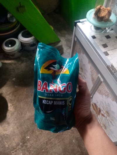 In Pompanua, Indonesia, a person's hand is holding a teal-colored pouch of Bango Kecap Manis, a sweet soy sauce, in front of a concrete floor. Behind the pouch, two white containers with black lids are visible, along with a bright green object leaning against them. To the right, a white counter with a tiled surface displays a light blue plate holding several round, brown cookies. The overall scene suggests a domestic or outdoor storage area.