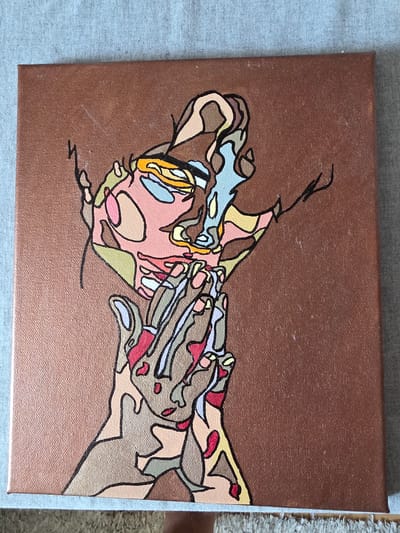 The image shows a square canvas painting with a dark brown background. The painting depicts an abstract figure composed of fragmented shapes and lines in various colors including peach, light brown, dark brown, yellow, light blue, and red. The figure appears to be a face and hands, rendered in a style reminiscent of cubism or stained glass, with bold black outlines separating the colored sections. The canvas is placed on a textured surface, likely a rug or textile, with a light blue and grey background visible at the top and bottom edges of the frame. There is no visible indication of its location in the city of Brzeg, Poland.