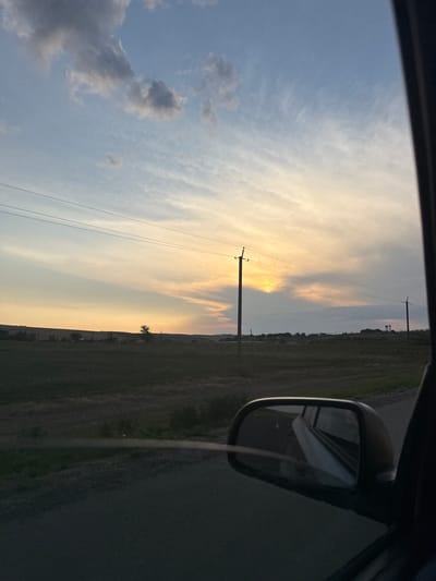 An exterior, medium shot captures a rural landscape at dusk, viewed from the perspective of a car. The sky is filled with a mix of dark and light clouds, with a gradient of pale yellow to blue indicating sunset. Several utility poles with wires stretch across the frame, bisecting the sky. In the foreground, a car's side mirror is visible on the right, reflecting a portion of the car's interior and the road ahead. The landscape beyond the road features a field of dry grass and scattered trees on a gently rolling hill. A single, solitary tree stands in the middle distance. The scene is set in the vicinity of Sadaclia, Moldova.