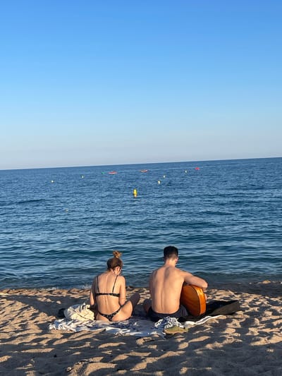 A man and a woman are sitting on a sandy beach in Canet De Mar, Spain, facing the ocean. The woman, on the left, is wearing a black bikini with a floral pattern and has her hair tied up in a bun. The man, on the right, is shirtless and wearing dark shorts. He is holding an acoustic guitar. They are sitting on towels on the sand, with a bag and a pair of shoes next to them. The ocean is blue and calm, with a clear sky above. In the distance, there are a few small boats and yellow buoys in the water. The sun is casting shadows on the sand.