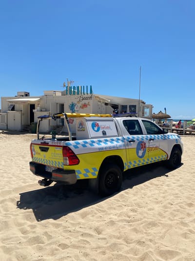 A Toyota Hilux pickup truck, marked with "Ambulancias Barbate," is parked on the sandy beach of Fuguilla Beach, in the city of Conil de la Frontera, Spain. The truck is white with yellow and blue checkered accents, and features medical symbols and phone numbers. In the background, a white building with "Fuguilla Beach" signage is visible, along with beach umbrellas, chairs, and a few people relaxing under them. The sky is clear and blue, and the overall scene depicts a sunny day at the beach.