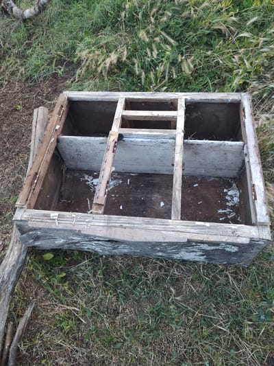 This media file is a photograph of a dilapidated wooden structure, resembling a shelf unit or cubbyhole, lying on the ground amidst overgrown grass and dry foliage. The structure appears old and weathered, with peeling paint and visible damage to the wood, suggesting it has been discarded or abandoned. It is divided into several compartments, some of which are open and empty, while others show remnants of old material, possibly paper or paint chips. The overall scene is outdoors, with a natural setting of vegetation and dry earth. There is no visible text in the image. The lighting suggests it might be daytime, but the specific time or weather conditions are not discernible. The location context provided is Kochmar, Bulgaria.