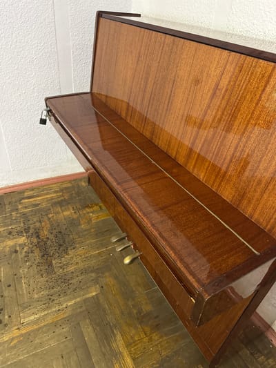 The image shows the keyboard and pedals of an upright piano. The piano has a polished wooden finish and is situated indoors, against a white textured wall. A small black padlock is attached to the edge of the piano. The floor is a parquet pattern with visible wear and discoloration. The scene is devoid of any people. The location is stated to be in Ракаўскі сельскі Савет, Belarus.