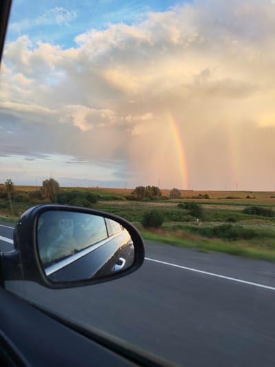 The image depicts a view from inside a moving vehicle, looking out towards a rural landscape and a double rainbow in the distance. The foreground shows the side mirror of the car, reflecting the interior and the sky. The side mirror is black and mounted on the car's frame. The reflection in the mirror shows a person holding a phone, suggesting the photo was taken by the passenger.
The road is visible in the bottom right portion of the image, appearing dark gray with a white line marking the lane. The landscape beyond the road consists of fields and scattered trees, indicative of a rural setting. In the distance, a prominent double rainbow stretches across a sky filled with dramatic clouds. The sky transitions from a clear blue at the top left to warmer, yellowish-orange hues towards the right, where the rainbow is located. The light suggests it is either sunrise or sunset.
There are several utility poles with wires visible in the distance across the fields. The location is identified as Bob'ya-Ucha, Russia. The overall scene captures a moment of travel through a scenic, rain-showered landscape.