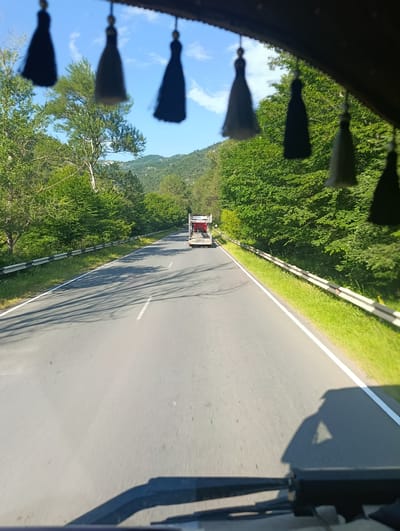 The image shows a view from inside a vehicle, looking forward through the windshield. In the foreground, the dashboard and part of the steering wheel are visible, along with the shadows of the vehicle's interior cast on the road. Suspended from the roof of the vehicle are several decorative dark tassels, which are slightly out of focus.

The main subject is a highway cutting through a lush, green, tree-lined landscape. The road is asphalt and has a white line marking the right edge. A guardrail runs along the right side of the road, separating it from a grassy verge. Further down the road, a truck with a white trailer is visible. The truck appears to be carrying some equipment, possibly construction materials or machinery. The road curves gently to the left as it recedes into the distance.

Beyond the trees, rolling hills are visible, covered in more greenery. The sky above is blue with a few scattered white clouds. The lighting suggests it is daytime, likely late morning or early afternoon, given the clear sky and the length of the shadows. The overall impression is one of a scenic drive through a natural landscape. The setting appears to be a rural or semi-rural area, given the abundance of trees and the relatively empty road. The specific location is Ijevan, Armenia. No people are directly visible in the image. The mood conveyed is peaceful and perhaps a sense of journey.