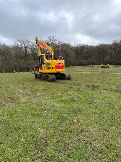 A construction site is depicted in a grassy field under an overcast sky in Telford, United Kingdom. A large yellow tracked hydraulic excavator, branded "H.E. Services," is centrally positioned, facing slightly right and away from the viewer. The excavator's tracks are dirty, and a person in high-visibility clothing is visible within its cab.
The field foreground and midground contain numerous pink triangular marker flags and yellow string lines laid out in a grid-like pattern on the patchy green and brown grass. Vehicle tracks are visible on the ground.
In the mid-to-far background, another piece of yellow machinery, possibly a drilling or piling rig, is active. Multiple individuals wearing high-visibility workwear (orange and yellow) are present in the background. One person is visible to the left near the distant tree line, while two to three others are near the drilling rig, performing ground-related tasks.
A dense line of bare deciduous trees forms the background, suggesting winter or early spring conditions. The overall scene indicates site preparation or surveying activities.