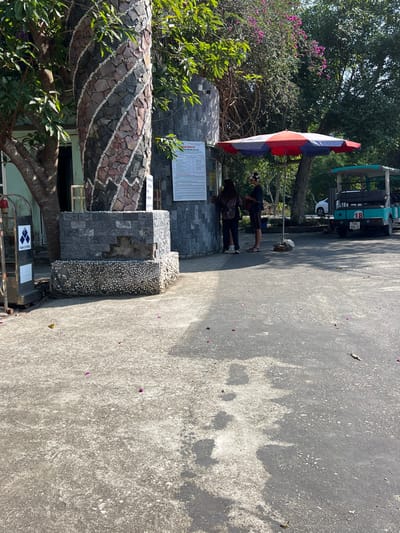 The image captures an outdoor scene, likely a pathway or entrance area in Trân Châu, Vietnam, under bright daylight. The primary subjects are two individuals, a decorative stone structure, and a large umbrella.
In the center-right, two people are standing. One individual, slightly taller, wears a dark shirt, shorts, and a patterned headband, facing towards the stone structure. The second person, slightly behind, is dressed in dark attire, also facing the structure. They appear to be observing or reading something. Behind them, partially obscured by trees, is a teal-colored electric shuttle or golf cart, marked with the number "18" and a visible Vietnamese license plate (e.g., "15B").
The left side of the image features a unique stone structure that resembles a pillar or wall. Its lower section is constructed from various dark, light gray, and reddish-brown stones arranged in a distinct wavy pattern, with bands of smaller, light-colored pebbles. The upper part is made of uniformly cut dark gray stones. A white sign is mounted on this structure. Above the structure and around it, lush green foliage, including trees and bushes with vibrant purple flowers, indicates a tropical environment. At the very bottom-left, a small black and white sign with an arrow and the text "TAY CONG" is visible.
The ground is a paved surface, likely concrete or asphalt, mottled with lighter and darker patches, suggesting wear or varying moisture levels. Strong sunlight casts clear shadows of the umbrella, people, and structures onto this surface. Scattered purple flower petals are visible on the ground.
The white sign on the stone structure clearly displays "NỘI QUY" at the top, which is Vietnamese for "REGULATIONS." Below this heading, the sign contains several lines of smaller, unreadable text, presumably outlining rules or information for visitors. The overall atmosphere is serene and sunny, characteristic of a pleasant day at an outdoor attraction.