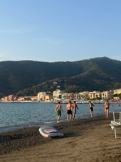 The image depicts a late afternoon beach scene in Sestri Levante, Italy, with the town's pastel-colored buildings and forested mountains under a clear sky. In the foreground, a dark sand and pebble beach hosts a deflated grey inflatable raft with a colorful design. Five individuals walk along the waterline from left to right: four males in swim trunks are in the lead, one appearing to jog. They are followed by a female in a dark swimsuit, seen from behind, who has a visible tattoo on her upper back. The calm, dark blue sea contains indistinct swimmers. The background reveals a strip of multi-colored buildings lining the coast, backed by steep, tree-covered mountains with scattered homes and a communication tower on a distant ridge.