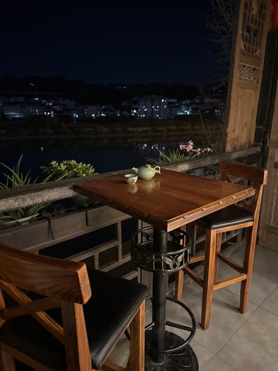 A high-angle shot depicts an outdoor setting in Zhuoshui, China, at night. The scene features a wooden table and chairs arranged on a porch. A tea set rests on the table, positioned near potted plants. The cityscape in the background is visible through the porch, illuminated by streetlights. The image captures a tranquil moment on the porch with an urban backdrop.