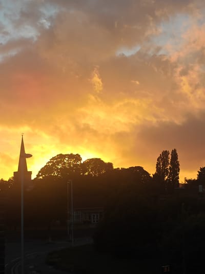 This media file captures a dramatic sunset or sunrise over a town or suburban landscape, likely in Worthing, United Kingdom. The sky dominates the upper two-thirds of the image, filled with rich, warm hues of intense orange, yellow, and reddish-brown, indicative of golden hour light. Clouds are heavily illuminated, creating a textured and vibrant canopy, with some cooler grey and pale blue tones visible in the higher, less directly lit areas.

The foreground and midground are largely silhouetted against this bright sky. On the left, a prominent church spire with a visible weather vane (a cross and an arrow) stands out as a dark shape. To its right, a dense band of trees forms a dark silhouette, with some taller, slender trees (possibly poplars) visible further right.

Beneath the tree line, a curving road or path is discernible, along with a few streetlights, all appearing as dark forms. A building, possibly a school, is partially visible through the trees in the mid-left, characterized by its windows and structure. Faint text spelling "SCHOOL" can be seen on a sign near this building. Another darker structure is visible further right. The overall impression is one of striking natural beauty, with the warm sky contrasting sharply with the shadowed land. No people are visible.