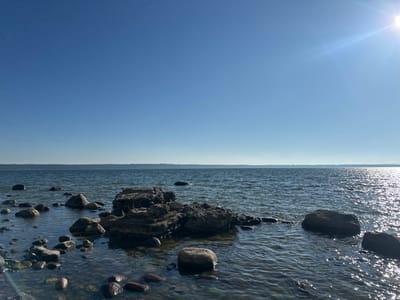 A body of water under a clear blue sky is seen from a rocky shoreline. The sun is shining brightly in the upper right corner, casting a glare on the water. The water is dark blue and rippled, with several large rocks protruding from it. The shoreline is made up of smaller pebbles and rocks. In the distance, a thin line of land is visible, with faint trees. The location is the coast of Tabasalu alevik, Estonia.